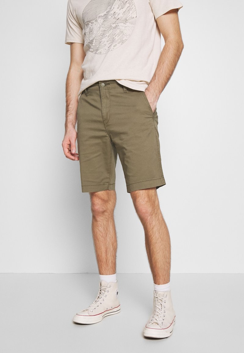 Shorts vert olive à coupe droite, en tissu de coton. Portés avec un t-shirt graphique beige et des baskets claires.