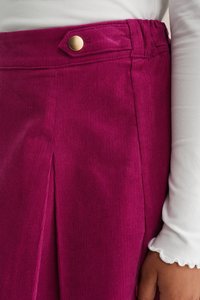 Fuchsia kordfløjlskørt med en guldknap i taljen, sidelæg-detalje og ribbet tekstur. Langærmet hvid bluse synlig.