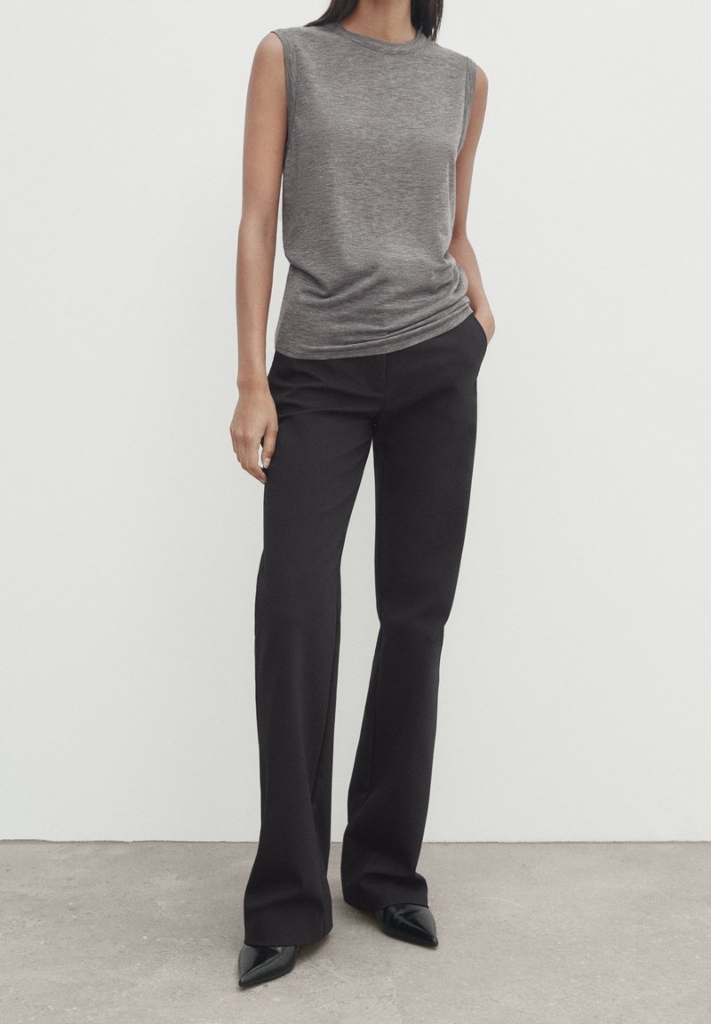 Graues ärmelloses Oberteil mit Rundhalsausschnitt, kombiniert mit schwarzen weit geschnittenen Hosen und spitzen schwarzen Schuhen, vor einem neutralen Hintergrund.