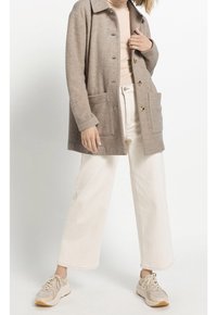 Cappotto in misto lana beige con vestibilità rilassata, due tasche frontali e bottoni beige, abbinato a pantaloni a gamba larga bianchi e sneakers beige.