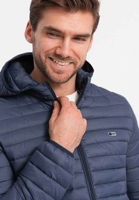 Giacca isolante blu navy con cappuccio, caratterizzata da una texture trapuntata, zip frontale e una toppa con logo sul lato sinistro del petto.