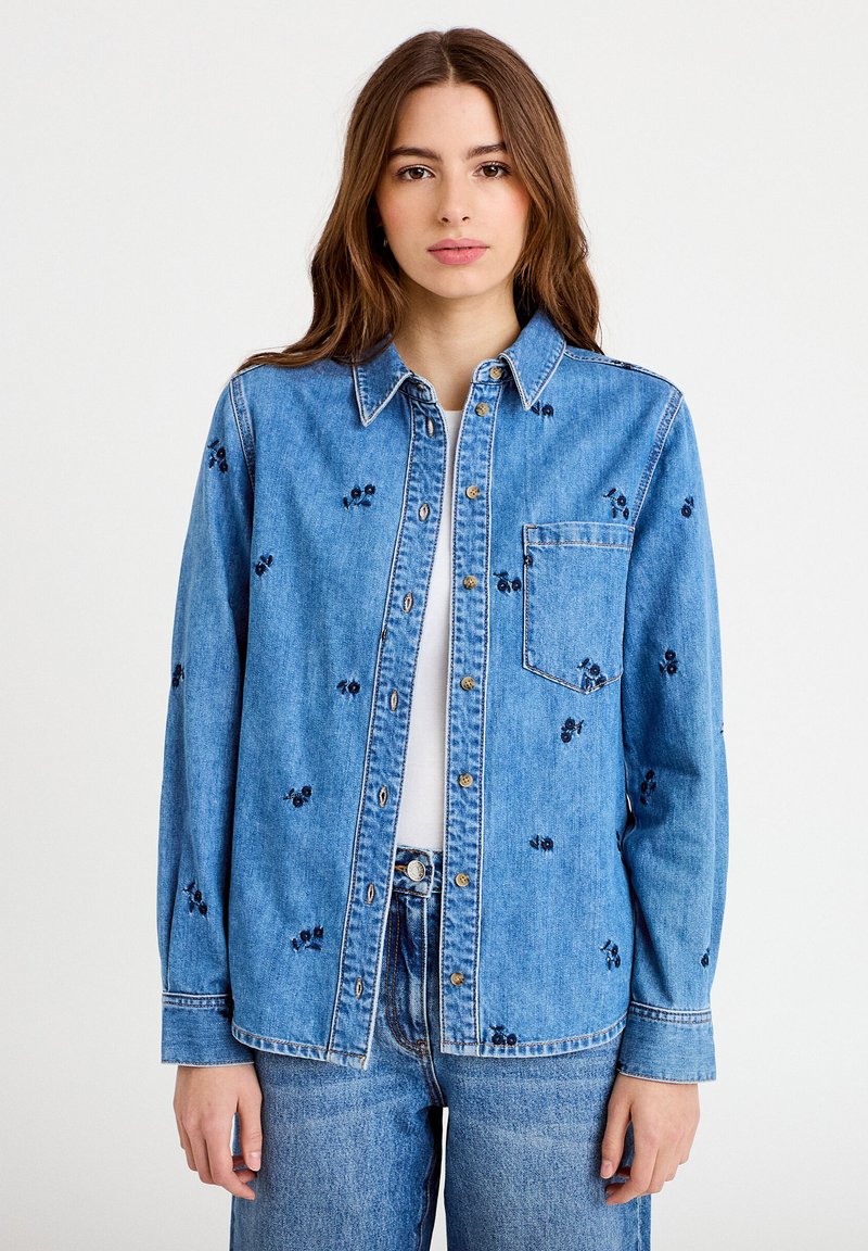 Ung kvinde iført en blå denim skjorte med knapper med små mørke broderede kirsebær over en hvid top og matchende blå denim jeans.