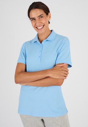 Lachende vrouw met donker haar draagt een lichtblauw poloshirt en grijze broek, staat met gekruiste armen tegen een effen achtergrond.