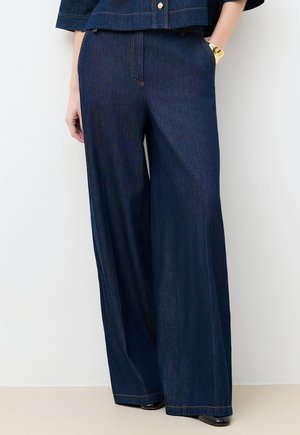 Szerokie, ciemnoniebieskie dżinsy noszone z dopasowaną kurtką, ręce w kieszeniach, na dole widoczne czarne buty.