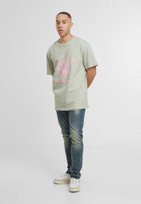 Zelené oversized tričko s růžovým grafickým potiskem, spárované s modrými džíny a bílými teniskami, na jednolitém pozadí.