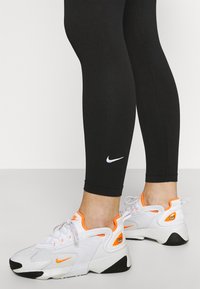 Vita sportskor i vitt med orange accenter, svart sula och intrikat textur; kombinerat med svarta leggings med en vit Nike-logotyp.