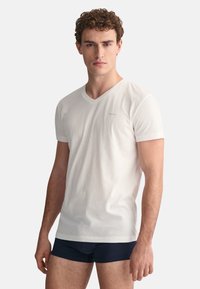 Weißes Baumwoll-T-Shirt mit kurzen Ärmeln und V-Ausschnitt, versehen mit kleinem Logo, getragen mit marineblauen Shorts. Einfaches Design, weicher Stoff und lässige Passform.