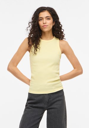 Femme aux cheveux bouclés foncés portant un haut jaune pâle sans manches et un pantalon noir, debout les mains dans le dos.