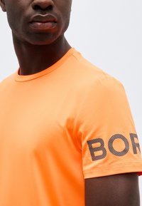 Άνδρας που φοράει φωτεινό πορτοκαλί κοντομάνικο πουκάμισο με μαύρα γράμματα με κουκκίδες "BOR" ορατά στο μανίκι, με απλό φωτεινό φόντο.