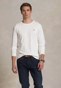 Homem jovem vestido com uma camisa de manga longa branca e jeans azul-escuro, de pé contra um fundo beige simples.