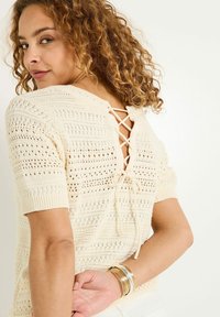 Top de punto crema con mangas cortas, que presenta un detalle de lazo en la espalda y un patrón de calados. Textura suave con bordes acanalados.