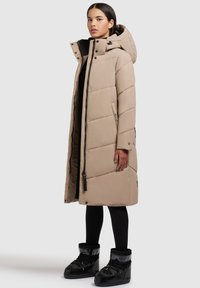 Beige Pufferjacke mit Kapuze, langer Schnitt, gestepptes Muster, Reißverschluss und seitlichen Taschen. Getragen mit schwarzen Stiefeln und Leggings.