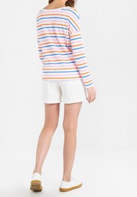 Camisa de manga larga a rayas en blanco, naranja, azul y rosa, combinada con pantalones cortos blancos y zapatillas blancas, vista desde la parte trasera.