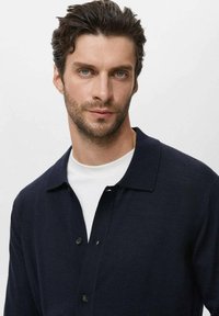 Hombre con cabello corto y rizado oscuro y barba clara, vistiendo un cárdigan negro con botones sobre una camisa blanca, fondo neutro.