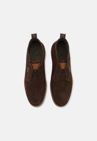 Scarpe in suede marrone con punta rotonda, design con lacci, suola in gomma e un'etichetta in pelle marchiata di lato. Linguetta di estrazione con dettagli rossi e neri.