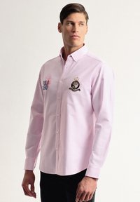 Camisa de botones de manga larga de color rosa claro con cuello, detalle de logo bordado y costuras en contraste en el pecho.