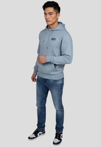 Felpa leggera blu con tasca frontale, cappuccio con lacci e testo nero. Abbinata a jeans blu e scarpe da ginnastica bianco e nere.