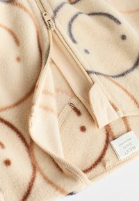 Fleece Babybekleidung in Creme mit hellbraunen und blauen Smiley-Gesichtern, ausgestattet mit einem beigen Reißverschluss und weichem, strukturiertem Stoff.