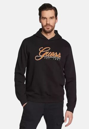 Man wearing black hoodie with "Guess Est. 1981" text standing with one hand in pocket against white background.