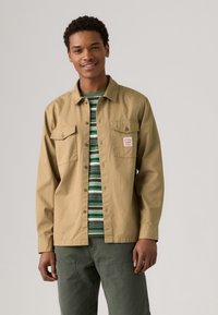 Lichtbeige overhemd met knopen en twee borstzakken. Daaronder een groen gestreept t-shirt. Model draagt een olijfgroene broek.