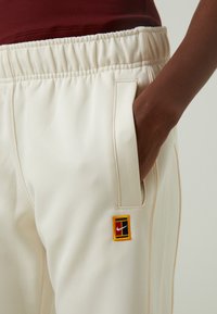 Pantalones deportivos de color crema con cintura elástica, bolsillo lateral y un parche de logo de Nike multicolor en la pierna. Textura suave.