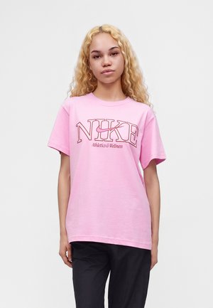 Jeune femme aux longs cheveux blonds bouclés, vêtue d'un t-shirt rose Nike et d'un pantalon noir, devant un fond blanc uni.