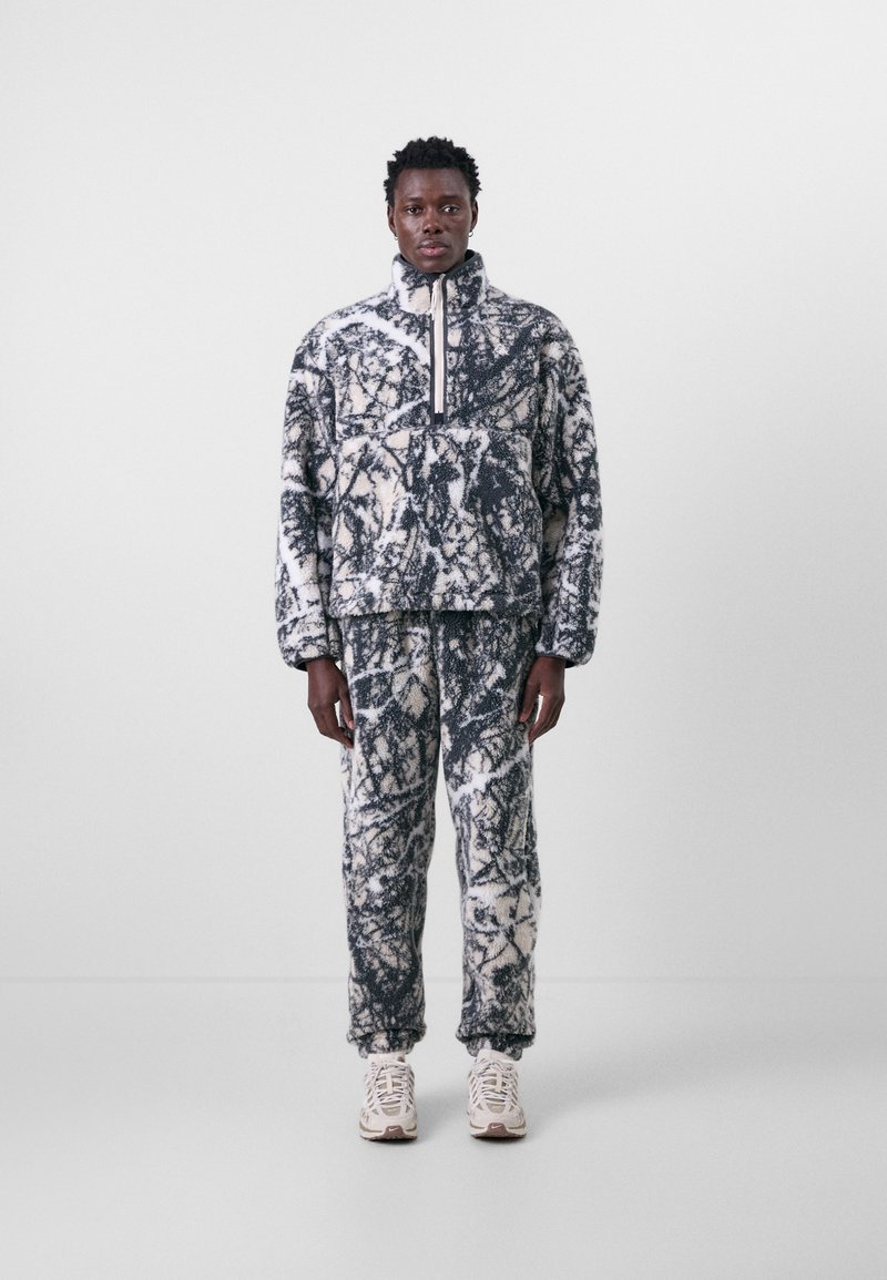 Fleece pullover and pants set with a black and white abstract pattern, featuring a high collar, zipper, and textured fabric. Paired with light sneakers.