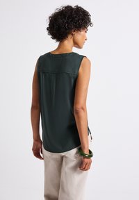 Bezramieniowy ciemnozielony top o miękkiej fakturze, z plecowym yoke i bocznymi rozcięciami. Zestawiony z jasnobeżowymi spodniami i zieloną bransoletką.