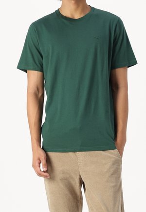 Mand iført en ensfarvet mørkegrøn kortærmet t-shirt og beige fløjlsbukser med den ene hånd i lommen mod en hvid baggrund.