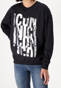 Sudadera negra con una gran impresión plateada desgastada que dice "ICU" e "ICON", con cuello redondo y puños acanalados. Combinada con jeans de mezclilla.