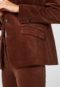 Blazer in corda di colore marrone con rever a punta, due bottoni e una tasca laterale. La texture è a coste, aggiungendo profondità al tessuto.