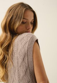 Jasnobrązowy bezrękawnik z dzianiny, z wzorem w warkocze, ściągaczem przy szyi i fakturowanym wykończeniem, noszony z luźnymi falującymi włosami.