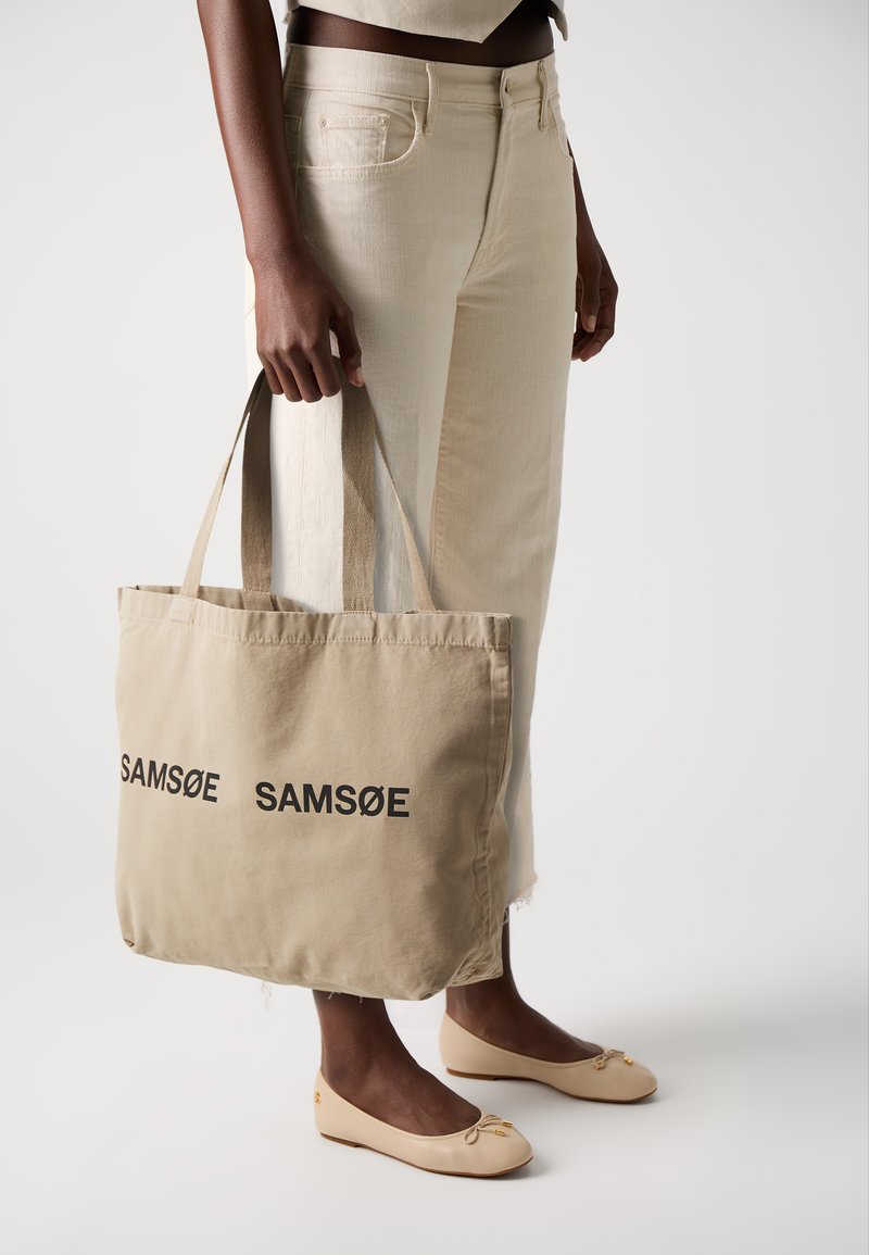 Beige Baumwoll-Umhängetasche mit zwei Griffen, auf der eine schwarze Aufschrift "SAMSONE" aufgedruckt ist. Getragen mit hellen Hosen und Ballerinas.