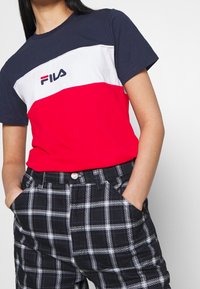 Rood, wit en marineblauw kleurgebroken T-shirt met korte mouwen en "Fila" logo. Gecombineerd met zwart-witte geruite broek en een knoopsluiting in de taille.