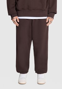 Brązowe dresowe spodnie z polarowego materiału mają elastanowy pas i mankiety, w zestawie z białymi sneakersami. Gładka faktura, luźny krój i brak widocznych wzorów.
