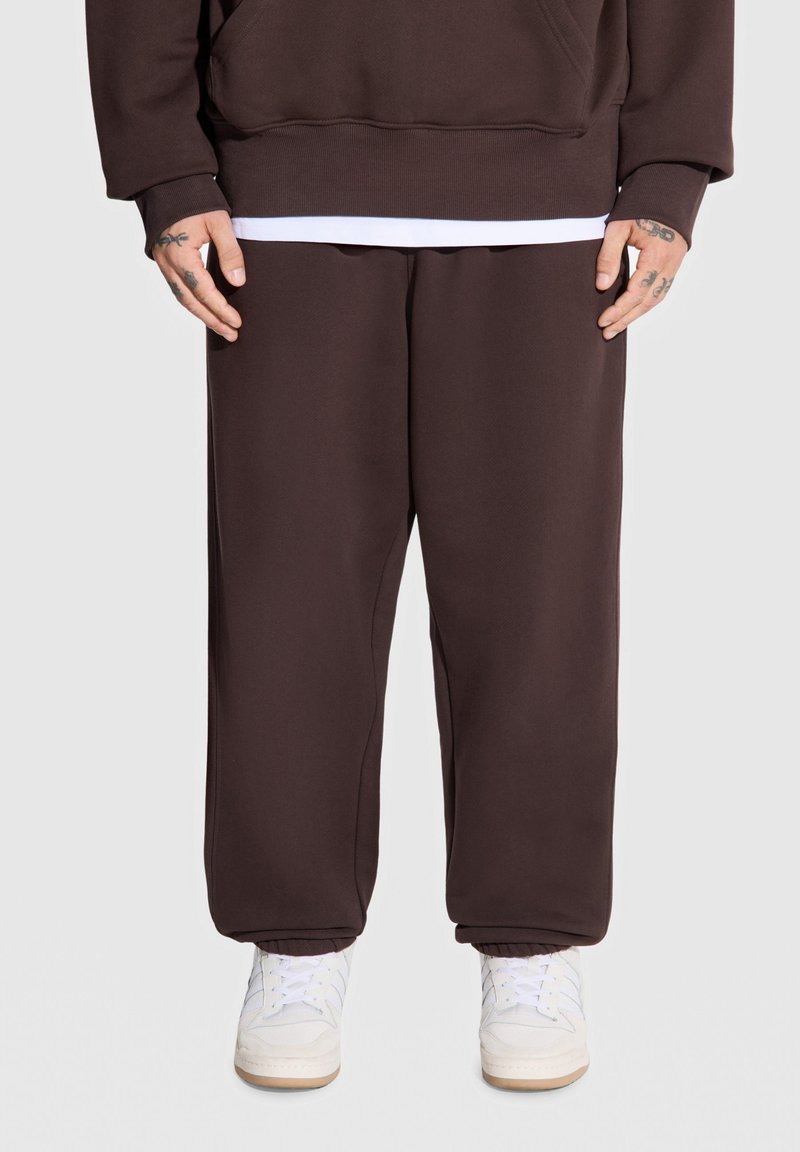 Brązowe dresowe spodnie z polarowego materiału mają elastanowy pas i mankiety, w zestawie z białymi sneakersami. Gładka faktura, luźny krój i brak widocznych wzorów.