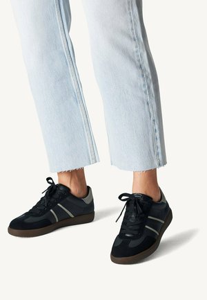 Osoba nosząca jasnoniebieskie jeansy z denimu i czarne trampki z szarymi paskami oraz brązowymi podeszwami na białym tle