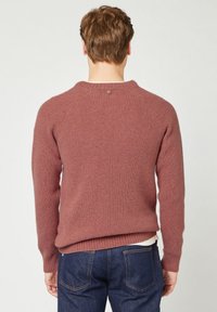 Mann mit kurz braunem Haar, der einen staubig rosafarbenen gerippten Pullover und dunkelblaue Jeans trägt, abgewandt vor einem einfarbigen hellen Hintergrund.