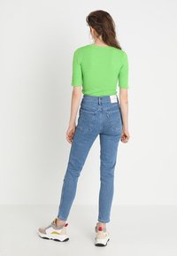 Top verde acanalado de manga corta combinado con unos vaqueros azules de cintura alta. Modelo con zapatillas beige con acentos de colores, de espaldas.
