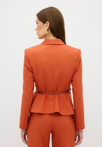 Oranje op maat gemaakte blazer met een peplumvorm, voorzien van een bijpassende riem in de taille en een gelapte kraag. Glad materiaal met een gestructureerde silhouet.