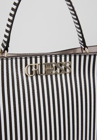 Sac à main à rayures verticales noires et blanches avec logo GUESS doré et poignées assorties rayées sur un fond uni.