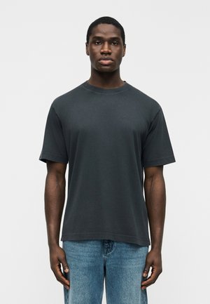 Młody mężczyzna stojący przodem, ubrany w jednolity ciemnoszary t-shirt z krótkim rękawem i jasnoniebieskie dżinsy, na jasnym tle.