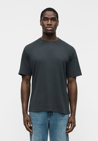Jeune homme debout face à l'avant portant un t-shirt à manches courtes gris foncé uni et un jean bleu clair sur un fond clair.