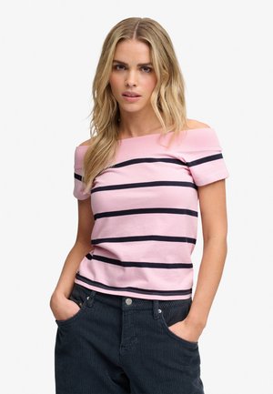 Lyshåret kvinde iført en pink off-shoulder top med sorte vandrette striber og mørkeblå manchesterbukser med hænderne i lommerne.