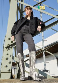 Botas brancas e pretas até ao joelho com um padrão texturizado, apresentando um salto bloco, combinadas com jeans escuros e uma jaqueta bomber preta.