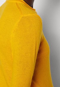 Pull en maille jaune, texture légère, col rond et détails côtelés au niveau des épaules. Vue en gros plan mettant en valeur le motif de la maille.