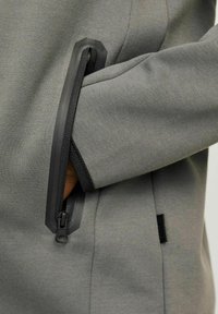 Felpa grigia con una texture liscia, dotata di una tasca con zip e hardware nero. Il design include una manica aderente e cuciture sottili.