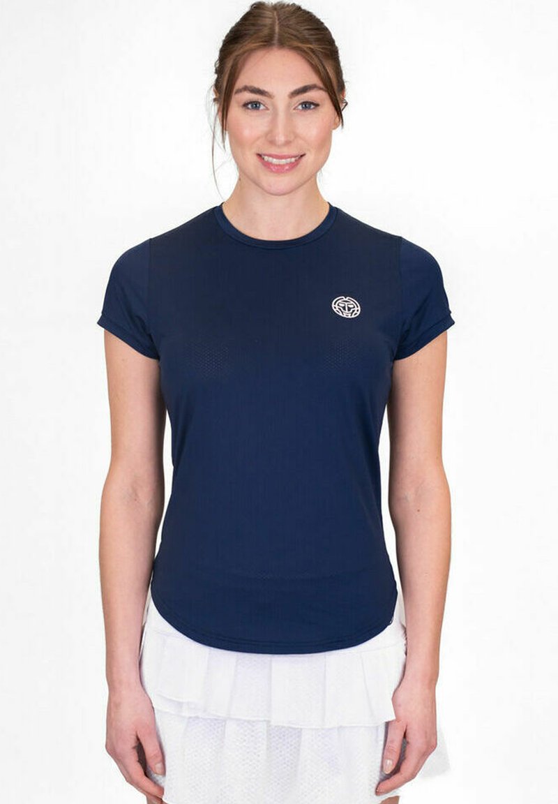 Donna con capelli castani che indossa una maglietta a maniche corte blu navy e una gonna bianca a strati, in piedi davanti a uno sfondo bianco semplice.