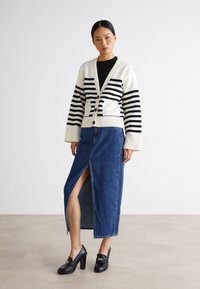 Mujer vestida con un cárdigan a rayas blancas y negras, blusa negra, falda de mezclilla azul con abertura frontal y mocasines negros de tacón, de pie sobre un suelo liso.