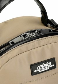 Sac à dos en nylon beige avec des accents noirs, doté d'une poignée supérieure, d'une fermeture éclair en métal et d'un patch logo rectangulaire sur le devant.